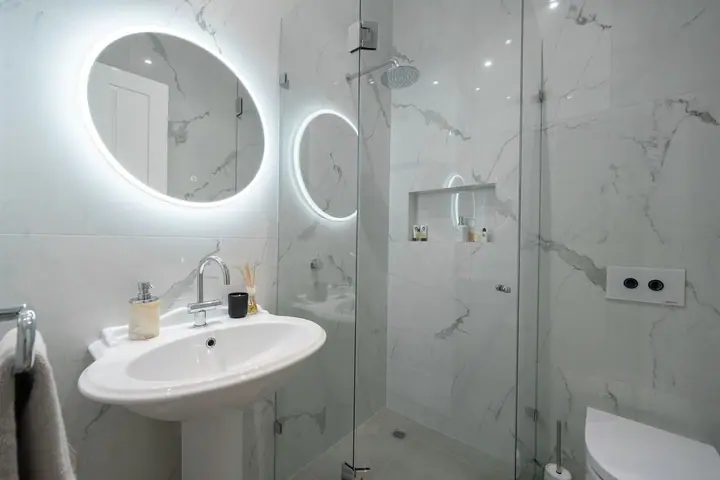 Backlit oval mirror, pedestal basin and frameless shower in a marble-look bathroom